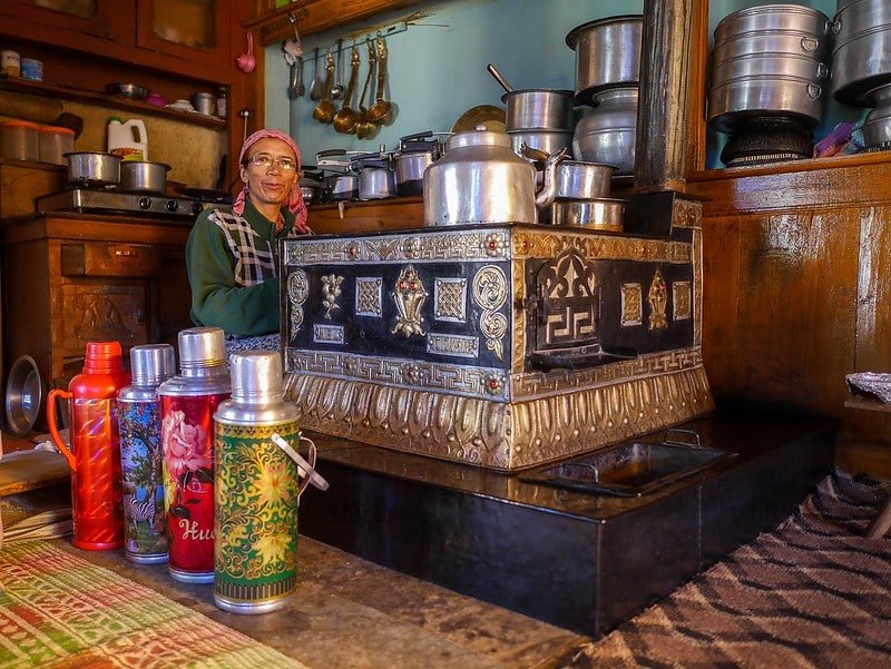 A woman in a traditional kitchen with colorful thermos flasks and metal cookware in ladakh