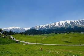 Scenic view of green meadows, winding road, and snow-covered mountains under a clear blue sky