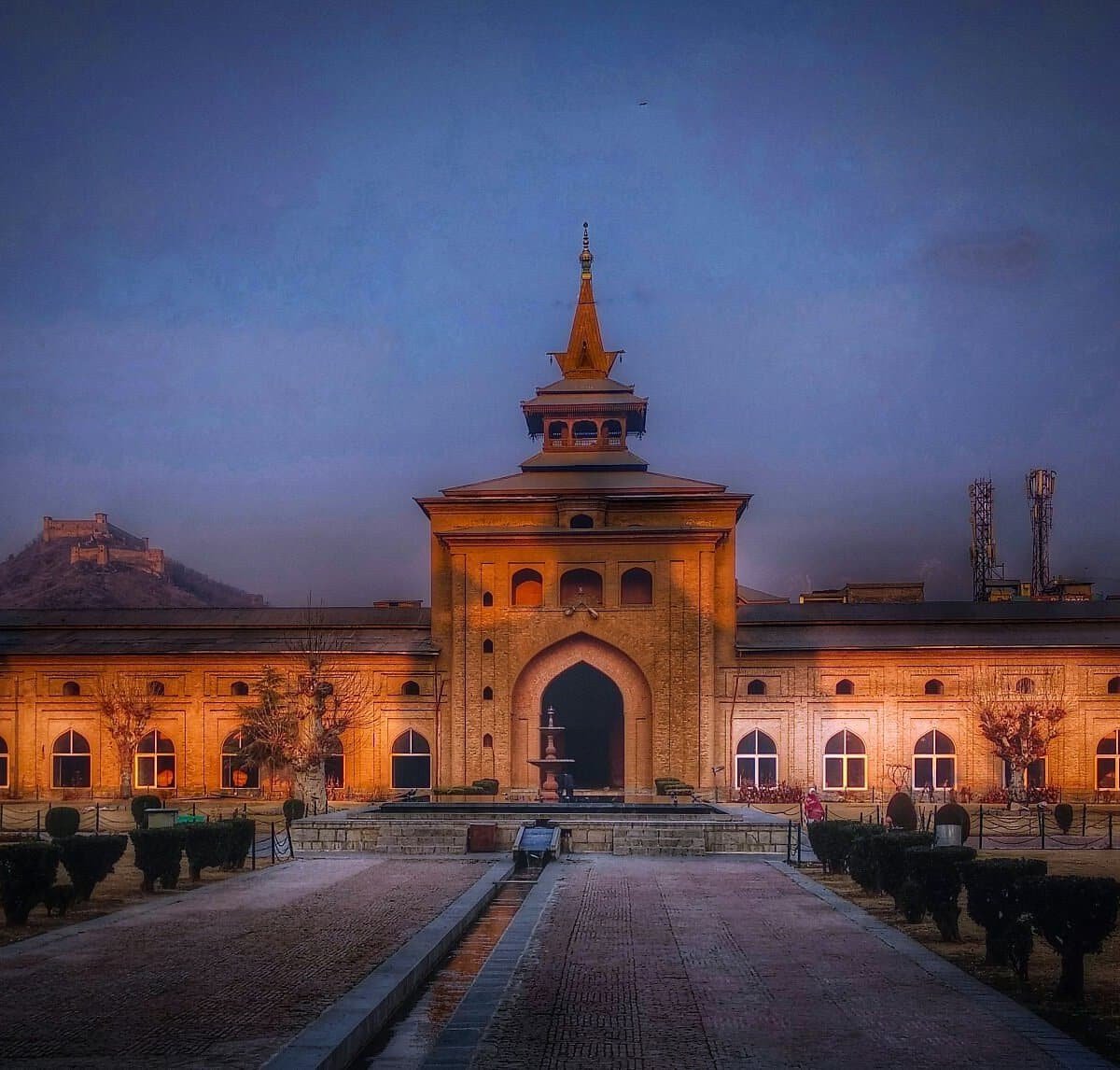 The Jamia Masjid in Srinagar at sunset, with golden light on its arched entrance and a distant fort on a hill behind