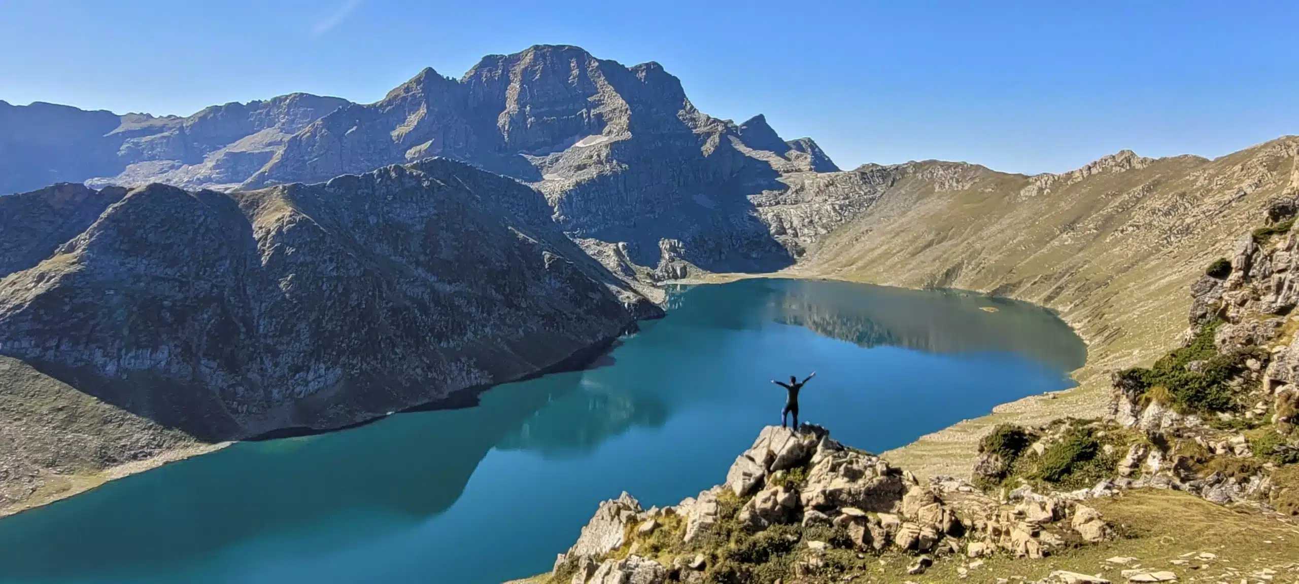 Trekker standing on a rock overlooking a deep blue alpine lake and majestic mountains on the Kashmir Great Lakes Trek