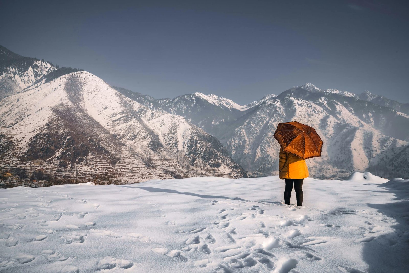 Person in a yellow coat holding an umbrella, standing on snow-covered ground with mountain ranges in the background