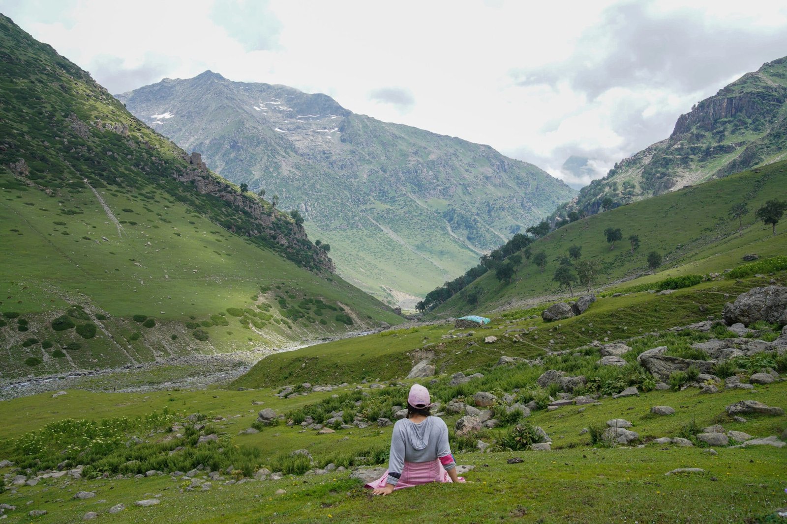 A person sits peacefully in a lush green valley surrounded by majestic mountains, enjoying the serene landscape and cloudy sky on here kashmir trip