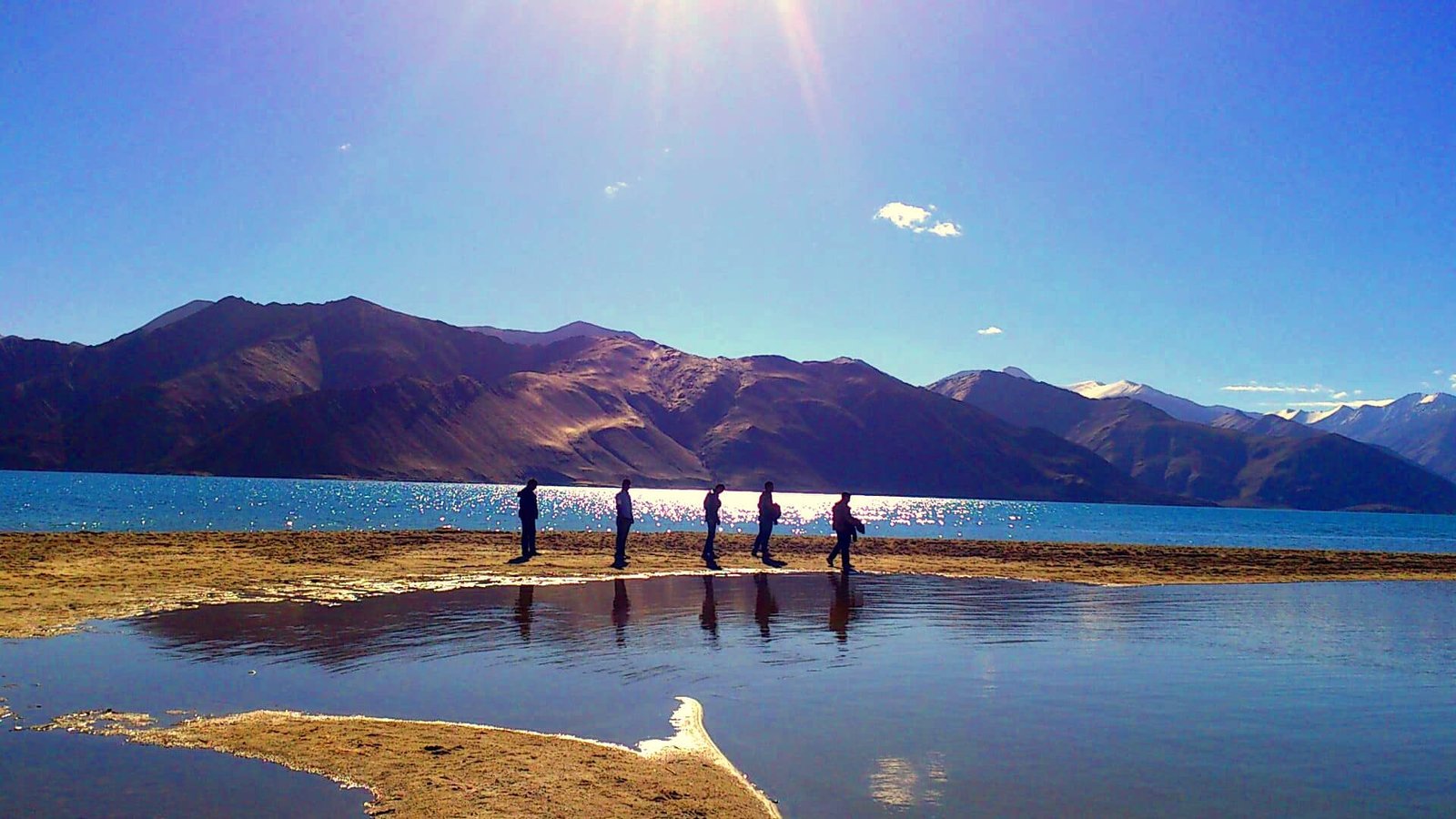 Silhouettes of people walking along the sandy edge of Pangong Lake with sparkling blue water