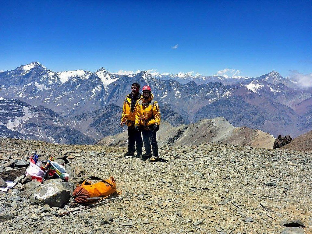 Two mountain climbers in yellow jackets and helmets stand on a rocky peak