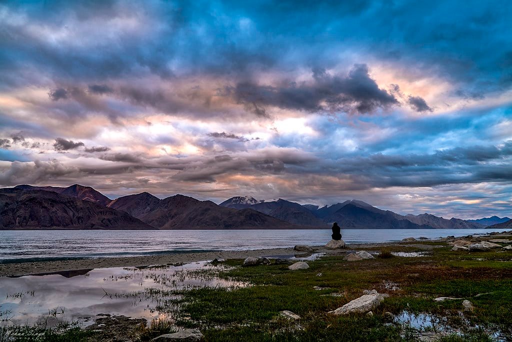 Person sitting on a rock by Pangong Lake under a dramatic, colorful evening sky with clouds