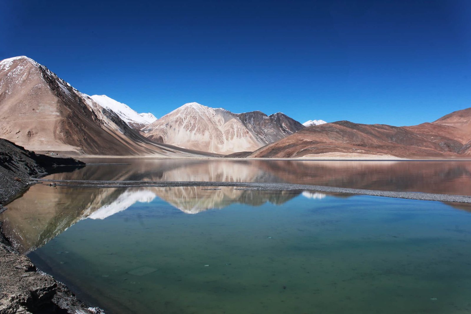 Clear reflection of snow-capped and brown mountains in the calm blue-green waters of Pangong Lake under a deep blue sky