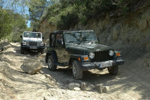 Two Jeeps off-roading on a rocky, uneven dirt path surrounded by vegetation and steep slopes.