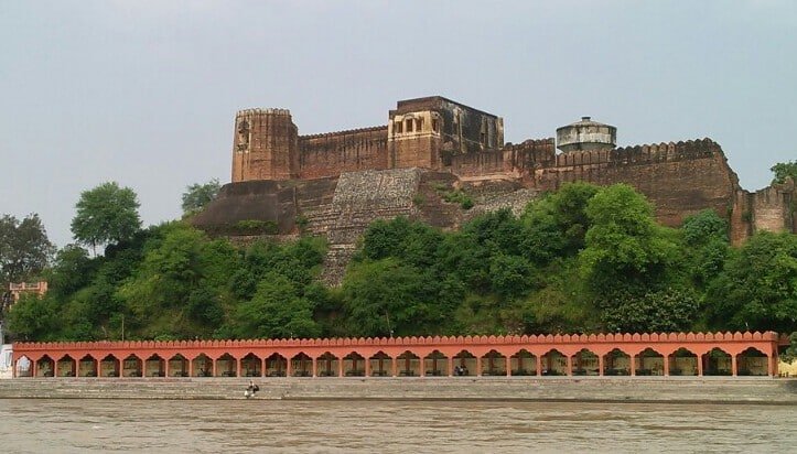 Akhnoor Fort on a hilltop with trees below and a riverside arcade in front.