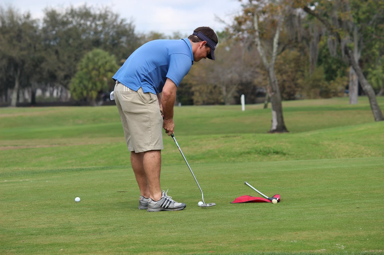 A golfer in a blue shirt and beige shorts lines up a putt on a green golf course with a flagstick and golf ball lying near the hole in gulmarg