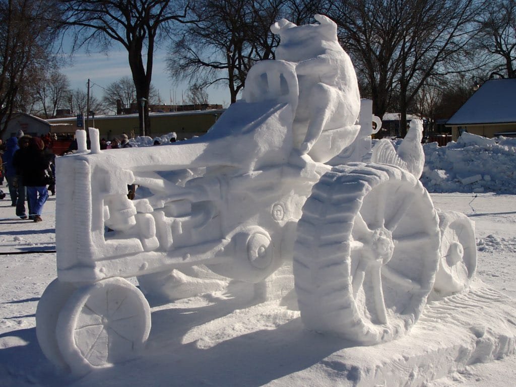 A detailed snow sculpture of a tractor with a pig sitting on the driver's seat and a chicken on the rear, displayed in an outdoor winter festival setting.
