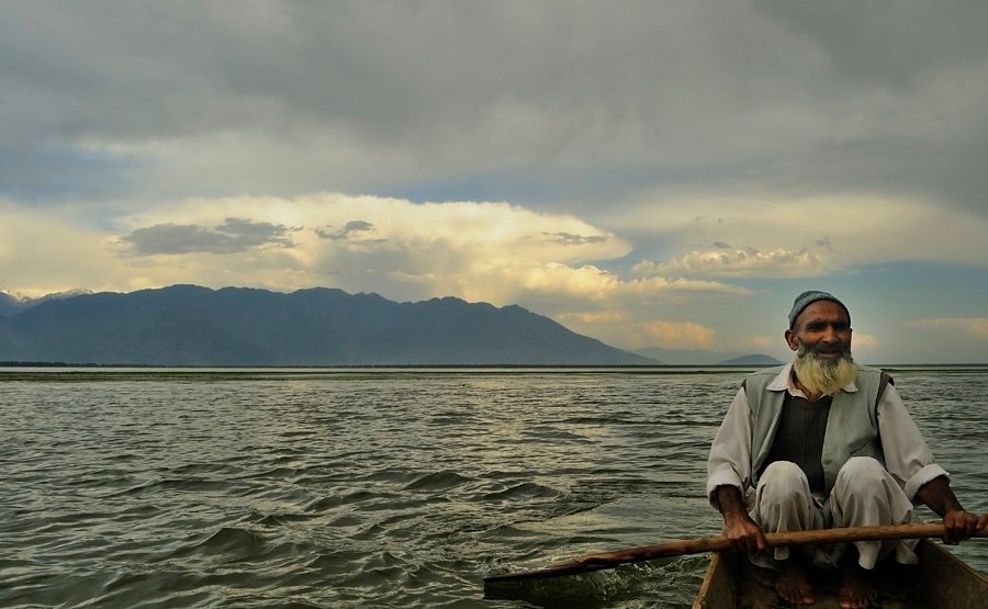 Elderly Kashmiri man rowing a boat on Wular Lake under a dramatic evening sky