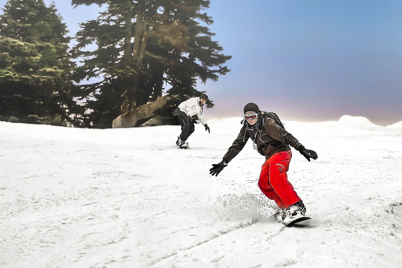 Two snowboarders ride down a snow-covered slope near pine trees under a clear blue sky