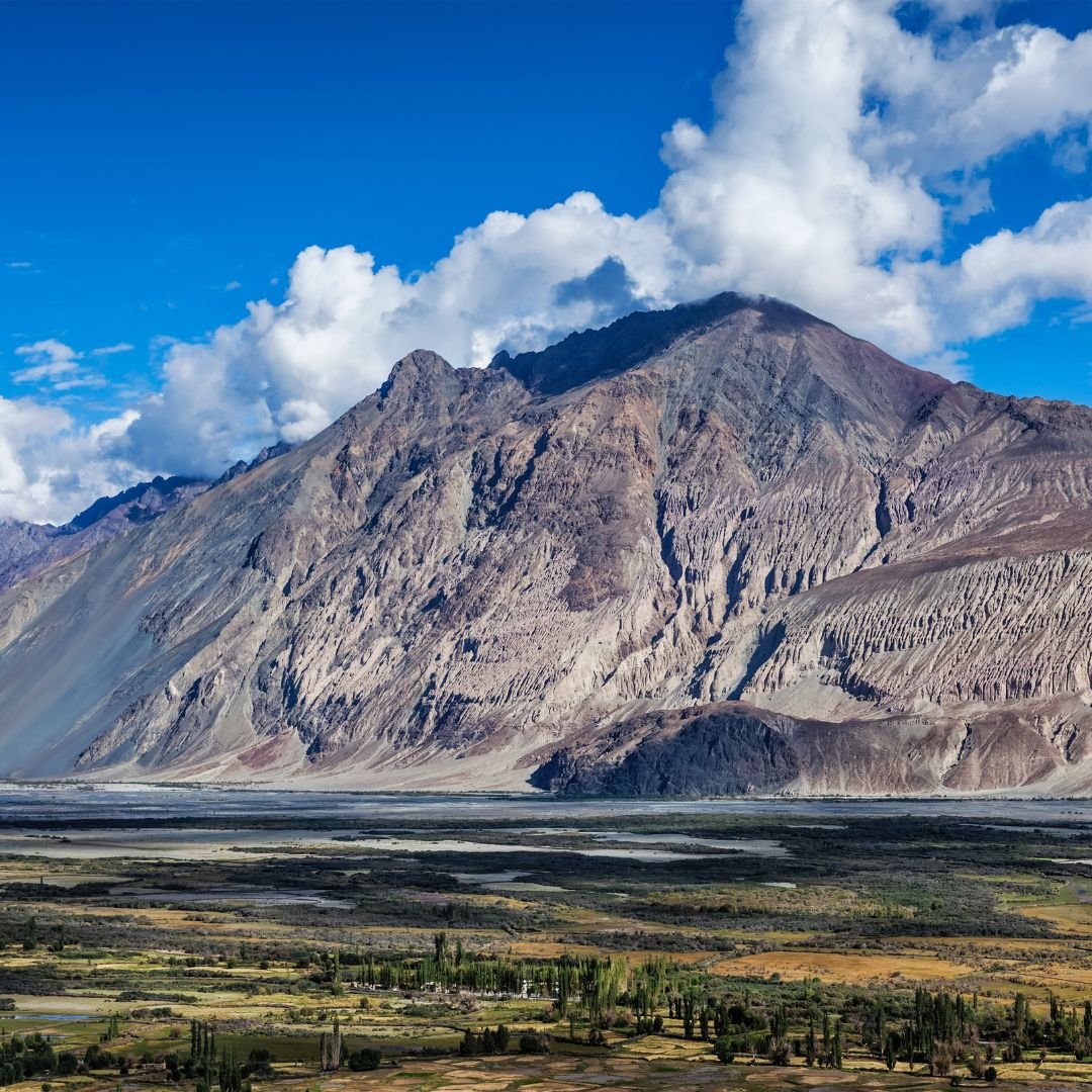 Massive rocky mountain near Siachen border with green valley fields under a partly cloudy blue sky