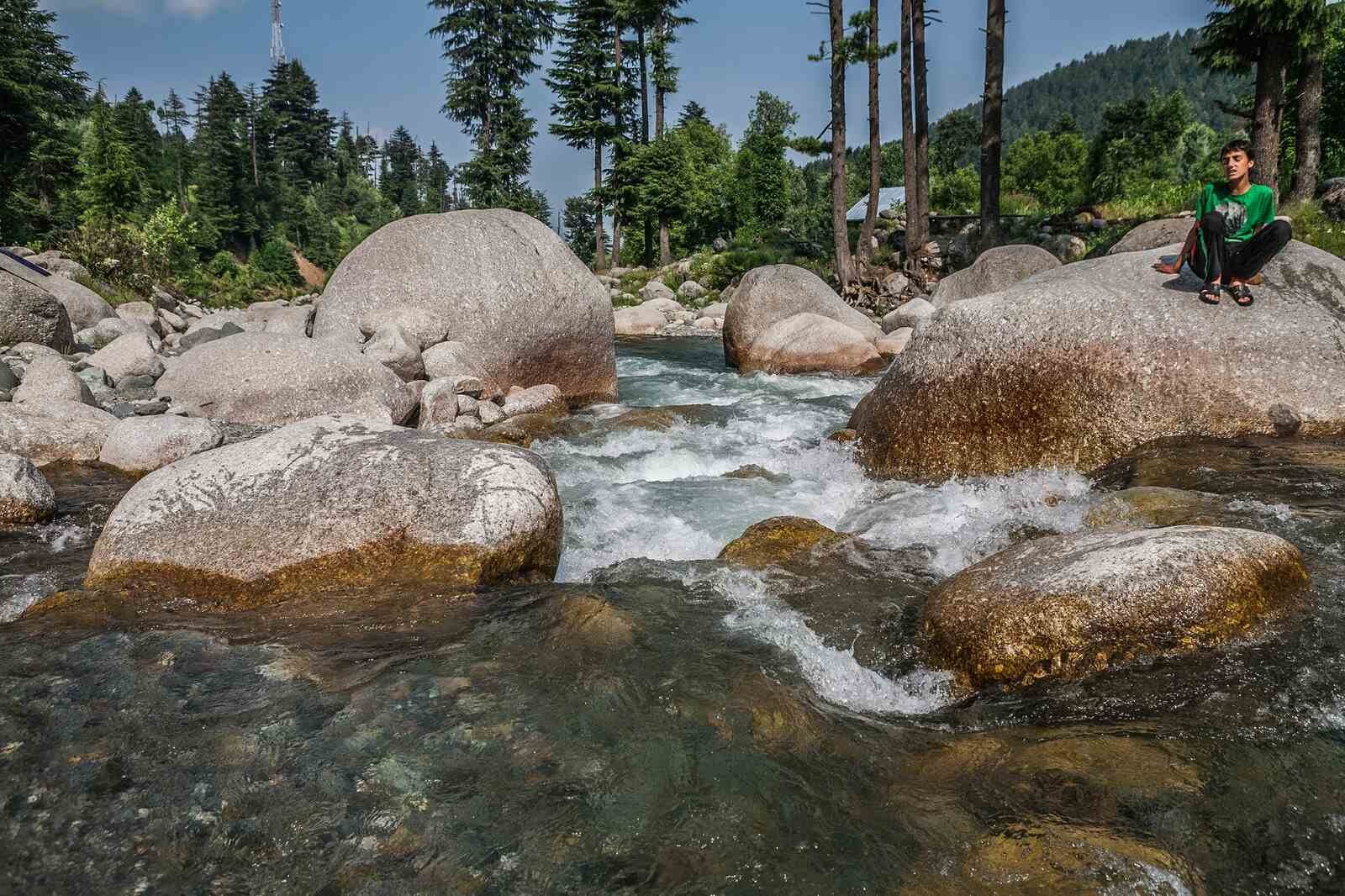Clear mountain stream flowing over rocks with pine trees and a boy sitting peacefully in Kashmir