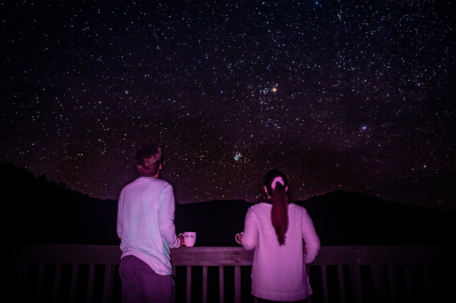 Two people stargazing from a balcony under a clear, star-filled night sky with mugs in hand