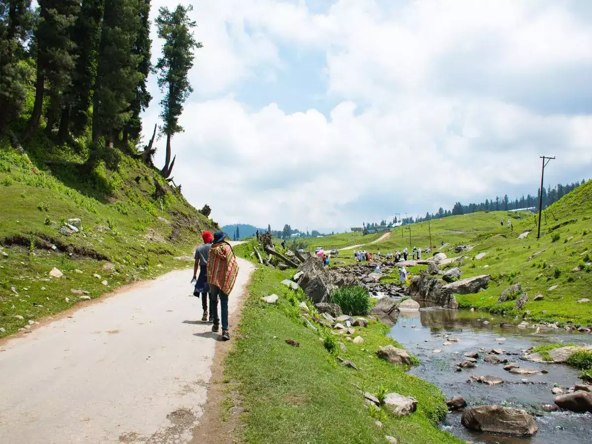 Tourists walking near a stream in Gulmarg during summer under clear skies and lush green surroundings